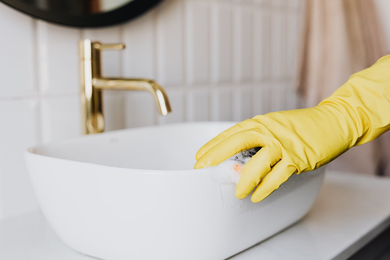 gallery-02 Crop faceless person in yellow latex protective glove cleaning surface of white stylish sink in contemporary bathroom interior